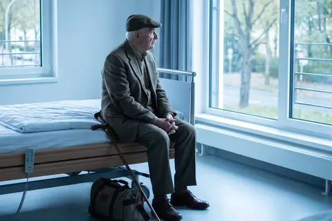 Symbolbild: Ein älterer Mann wartet mit gepackter Tasche auf der Bettkante im Klinikzimmer, KI-generiert